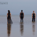 Three dancers facing front and back standing on the beach at low tide.