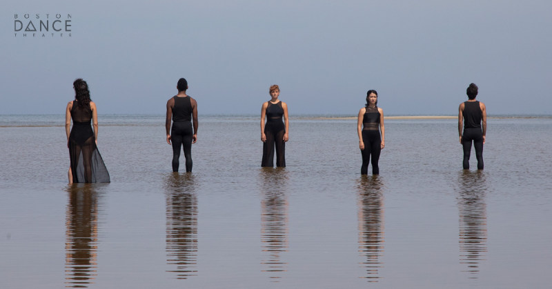 Three dancers facing front and back standing on the beach at low tide.
