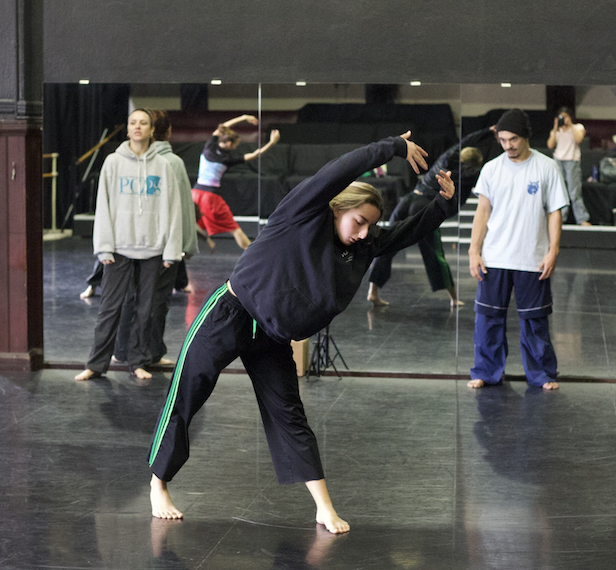 Choreographers Taylor Graham and Jerimy Rivera watch Trainee Addy as she poses with her arms raised in 5th position above her he
