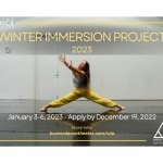 A dancer in yellow pants stands in a center split, reaching to the upper left corner of the image.