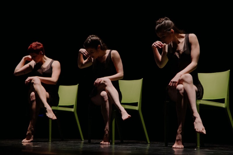 Three women each sitting on a chair with legs crossed and peering down to the side. 