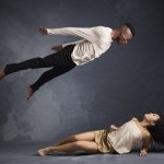 Photo depict female dancer on the floor looking up to her right as a male dancer is suspended in the air above her looking down 
