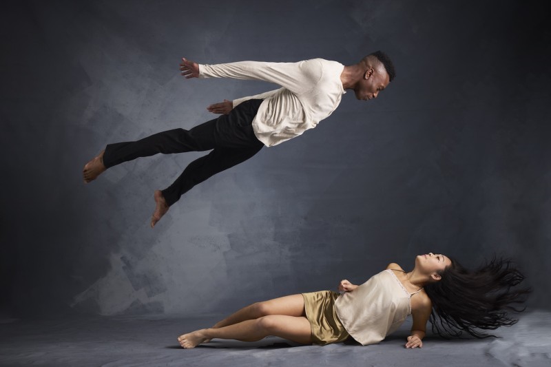 Photo depict female dancer on the floor looking up to her right as a male dancer is suspended in the air above her looking down 