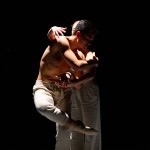 Image two dancers in a spotlight, one being hugged by the other.