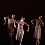 In front of an orange backdrop, a male dancer lifts another dancer high above his head