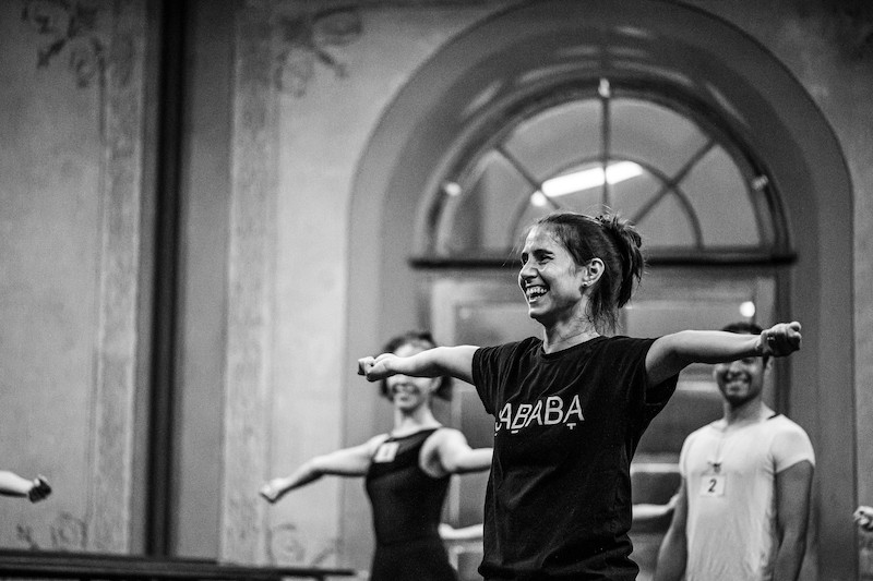A Black and White photo. Alejandra Jara is pictured in a black t-shirt. She is smiling with arms stretched wide to the side.