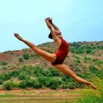 A dancer wearing a red leotard jumping above green grass in front of mountains.