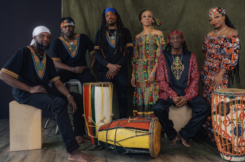 Image of Rasin Okan haitian roots musical ensemble, dressed in colorful printed patterns. Three drums are in the photo.
