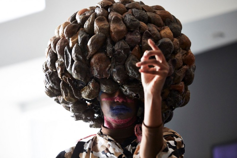 Black, female dancer with a large, bulbous wearable head sculpture, exhibiting terrestrial energy. Left hand raised to face. 
