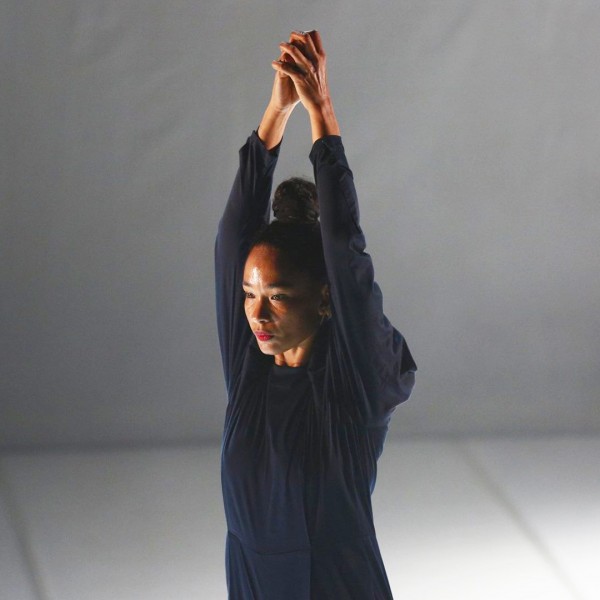 A woman with a bun, raises both arms with hands clasped in the air. Wearing an off-black, long sleeve jump suit.
