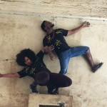 Two individuals, one crouched, on standing on one leg lock arms to form an architectural pose, against a backdrop of beige stone