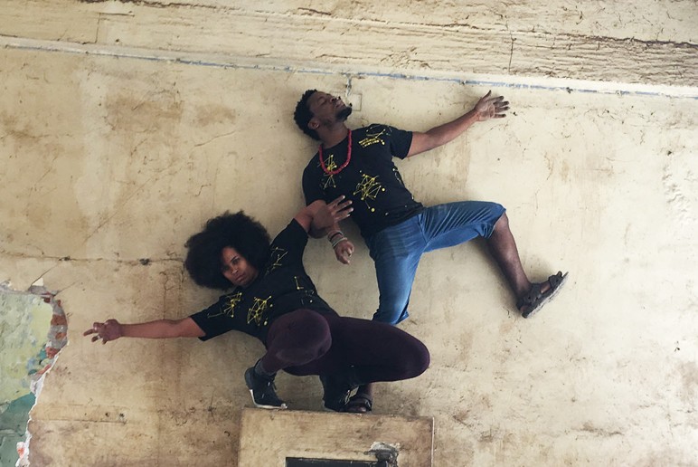 Two individuals, one crouched, on standing on one leg lock arms to form an architectural pose, against a backdrop of beige stone
