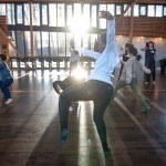 A dancer balances on one leg, sun shining through the studio windows.