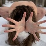 a naked woman crouches in the snow with her hands extended through her long brown hair. Her face is covered. 