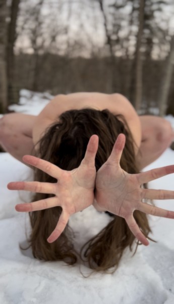 a naked woman crouches in the snow with her hands extended through her long brown hair. Her face is covered. 