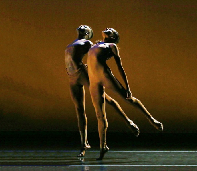 Picture of two dancers in brown unitards holding each other reaching for the sky