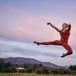 Male dancer in an orange unitard jumping outdoors with the sky behind him and his arms and legs stretched to his left side