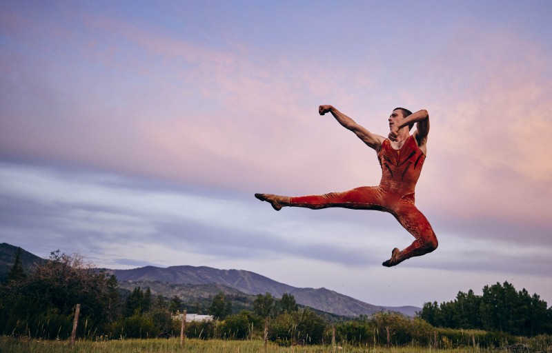 Male dancer in an orange unitard jumping outdoors with the sky behind him and his arms and legs stretched to his left side