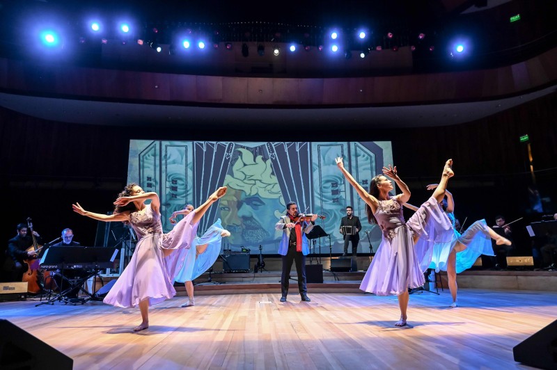 A group of dancers with violet sheer dresses and one leg extended with orchestra and violinist in the backdrop