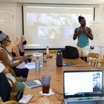 A group of people sitting around a table. One person presents in front of a projector screen with more people on Zoom.