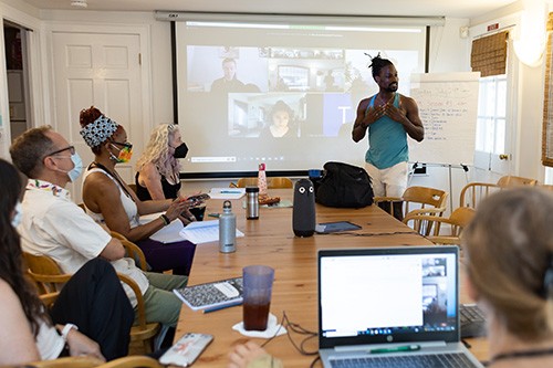 A group of people sitting around a table. One person presents in front of a projector screen with more people on Zoom.