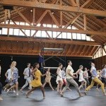 A group of dancers in a studio wearing assorted colorful street clothes run to the left. 