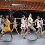 A group of dancers in a studio wearing assorted colorful street clothes run to the left. One dancer is posed on scaffolding in t