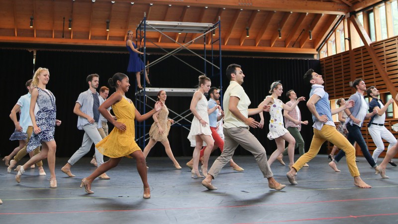 A group of dancers in a studio wearing assorted colorful street clothes run to the left. One dancer is posed on scaffolding in t