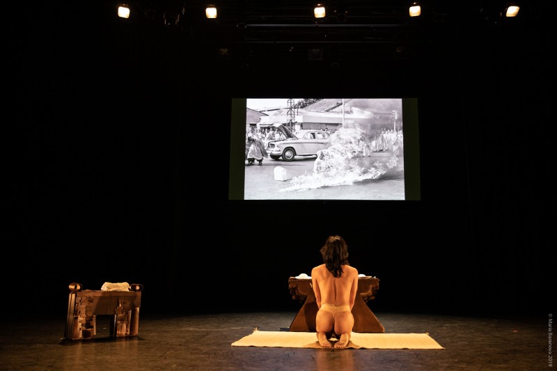 A half-naked performer kneeling in front of an altar. Projected on top is the image of Thich Quang Duc's self-immolation. 