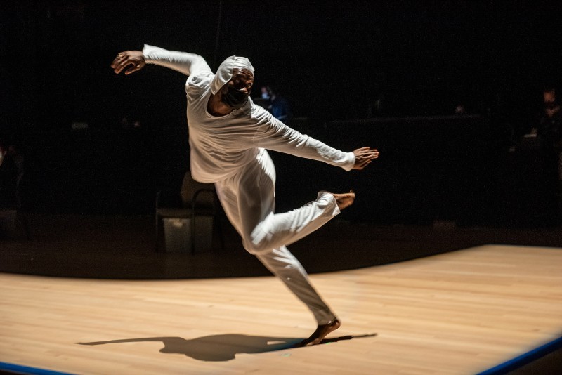 A single Black dancer wearing white is running forward looking over his left shoulder with his arms spread and left leg bent. 