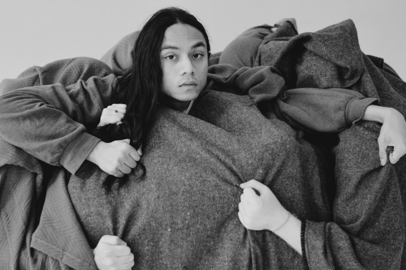 A person with long dark hair looks directly at the camera. Different hands reach at the blankets and at their hair.