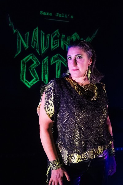 A woman with dark hair looks directly into the camera with Naughty Bits in green lettering behind her.