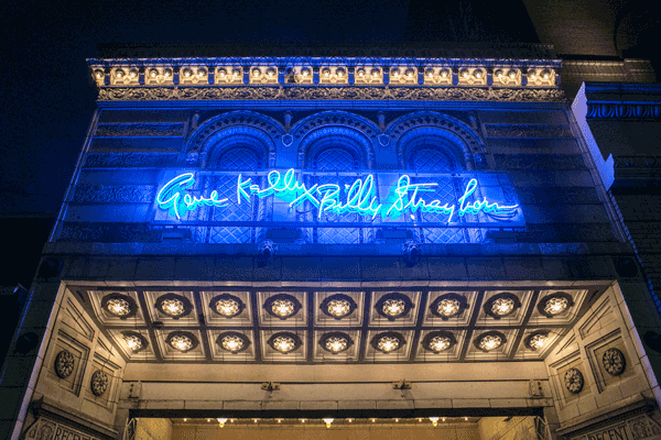 A photograph of an ornately tiled 1914 theater marquee with a blur neon sign that reads "Gene Kelly Billy Strayhorn"
