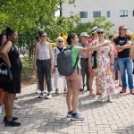 A group of people gather outside on a sidewalk. One person points away from the camera while the group looks and listens to her.