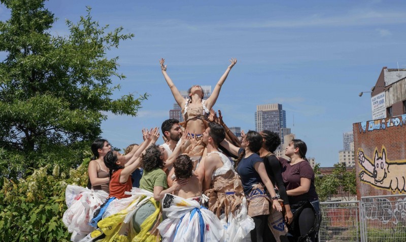 An image of a group of dancers wearing costumes made from recycled plastics, hold up a singular dancer in the air.