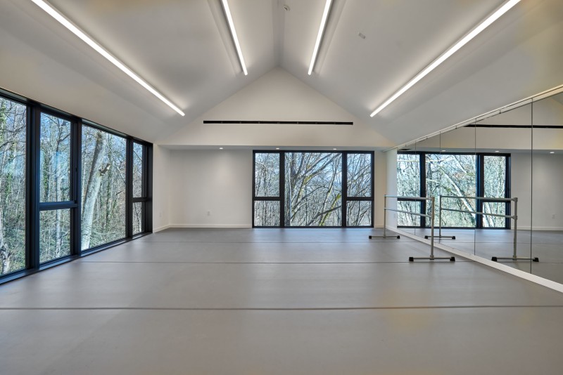 A brightly lit dance studio with grey marley floors, a mirrored walls and two large windows looking into a forest.
