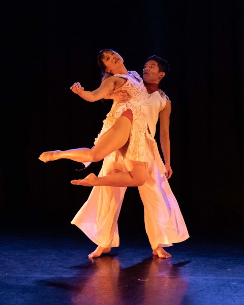 One dancer lifting another dancer dressed in white against a dark background