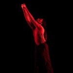 Ke'ron Wilson shining through red lighting on a dark stage, hands clasp and reaching up and out.