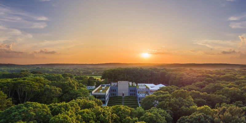 Bird's eye view of The Watermill Center, a large building surrounded by woods in front of an orange sunset. 