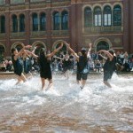  7 dancers wearing black shorts and tunics splash in a fountain, their arms joyously stretched overhead at the V & A. 