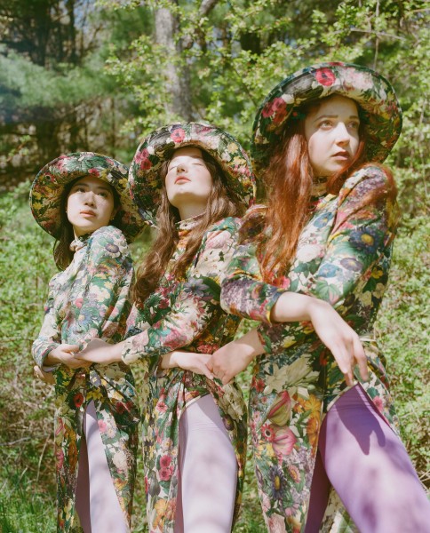 Three women stand shoulder to shoulder. They are wearing floral clothing in a lush outdoor setting.