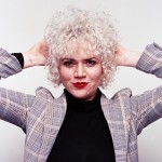 Woman with curly hair poses with arms on either side of her head