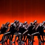 The dancers of Ballet Bc on stage with orange lighting. They are grouped together in a deep plie, wearing black bodysuits.