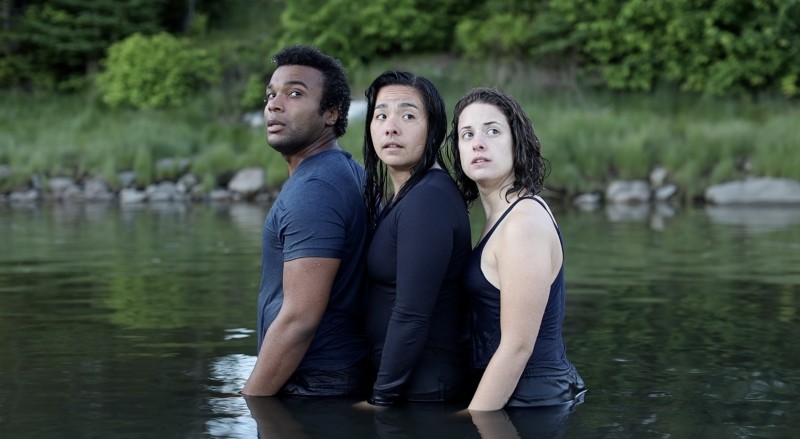 Three dancers stand close together, waist deep in the ocean with trees behind them, looking back with presence and hope
