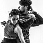 2 dancers move closely with one another with one looking left and the other looking forward. The photo is black and white.