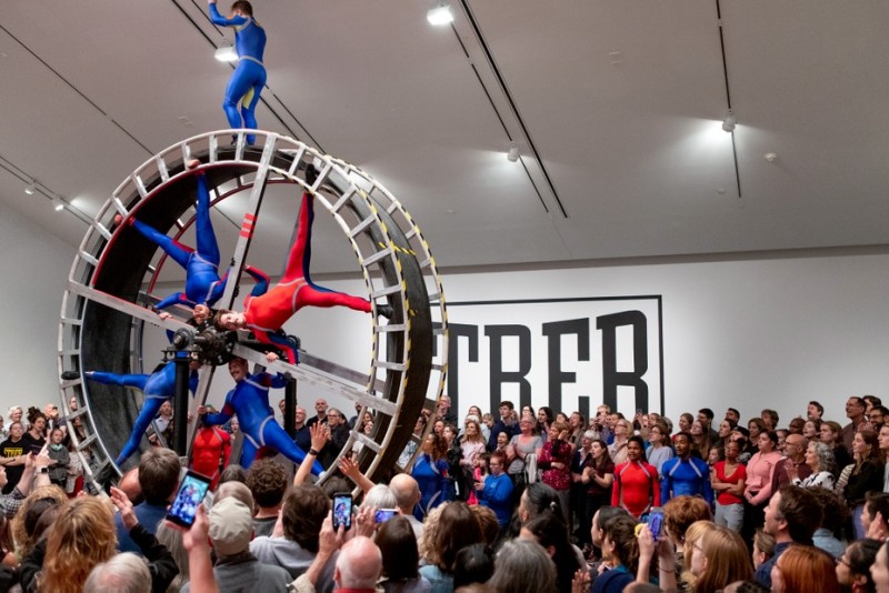 Dancers suspend in a giant wheel with large audience surrounding them