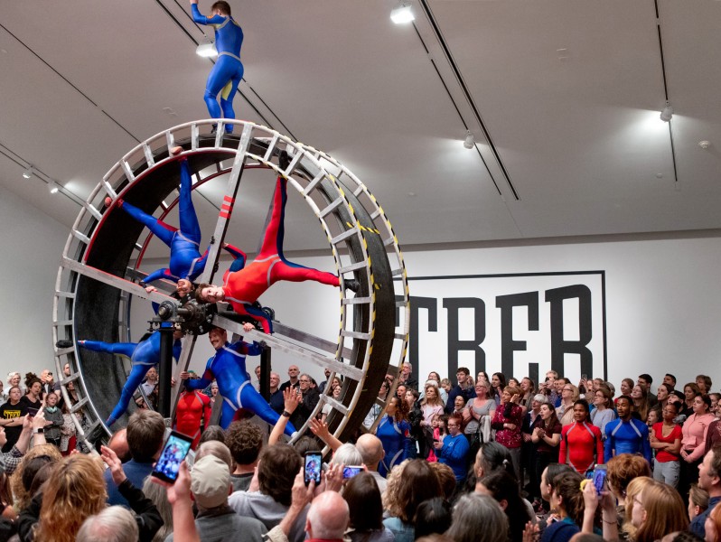 Dancers suspend in a giant wheel with large audience surrounding them