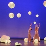 Dancers stand with arms raised overhead with a purple backdrop and illuminated lanterns surrounding them.