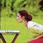 A woman in a white blouse and red pants sits at a table leaning forward while gazing into her laptop screen, sitting profile.