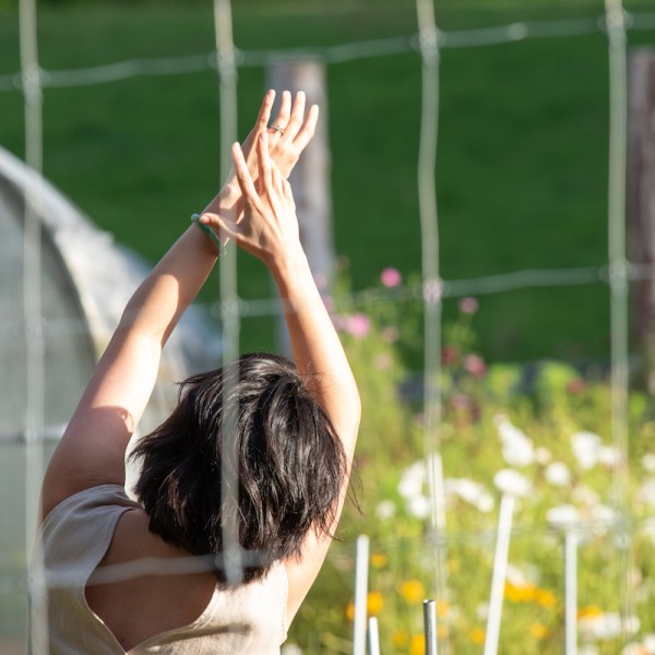 A woman stands in a garden with arms stretched overhead; she faces away from us and her black hair is short on her shoulders. 